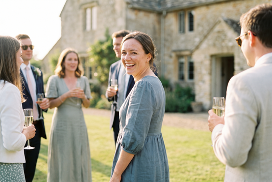 Wedding guests talking on a lawn outside a stone country house, one in a dusty blue midi dress