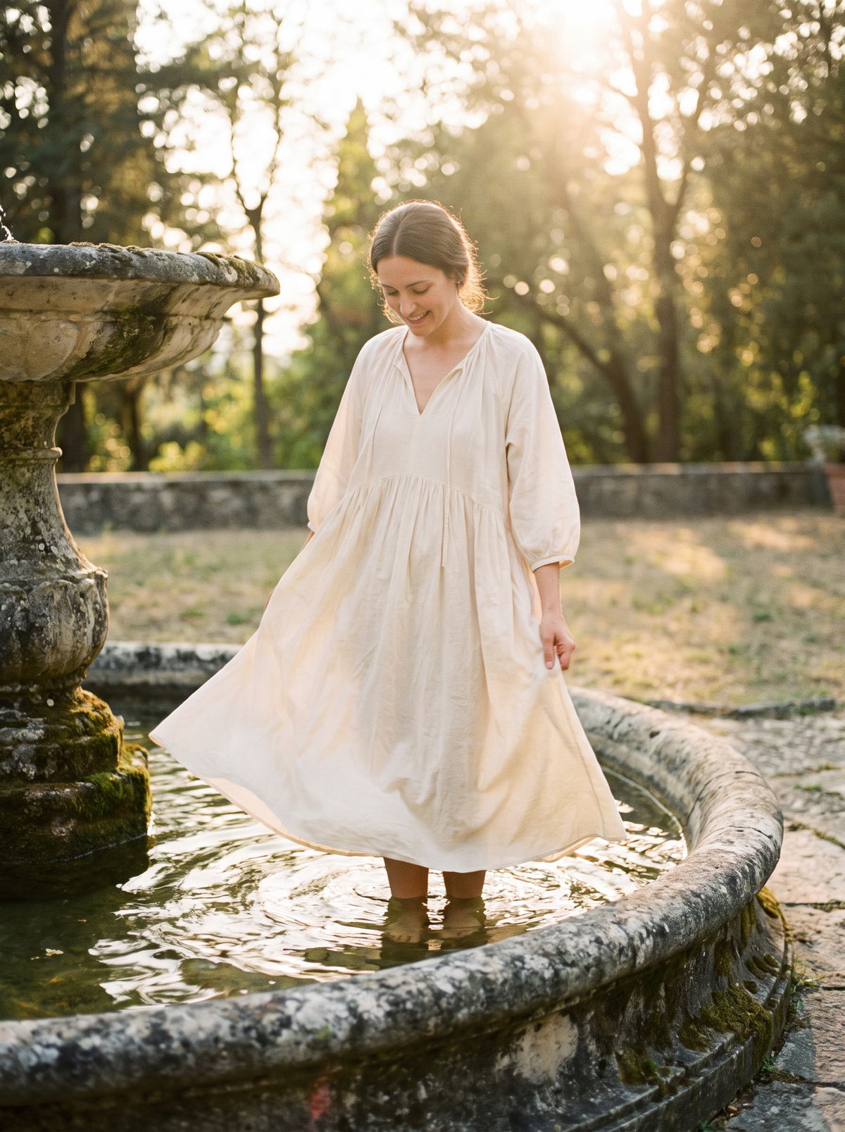 A woman standing in a shallow stone fountain in a long cotton dress